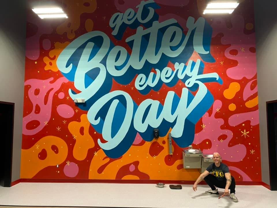 Jake crouching in front of his finished “Get Better Every Day” mural in bright red, pink and blue