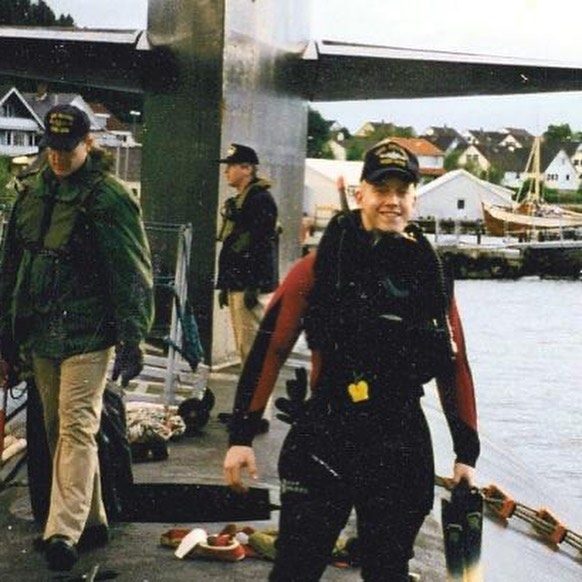 Jake in a wetsuit standing topside on a submarine with crewmates, a Norwegian harbor behind